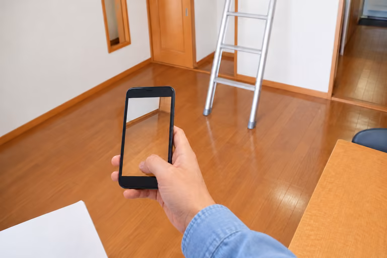 ロフトのはしごがある室内で、床と壁をスマートフォンで記録する手元