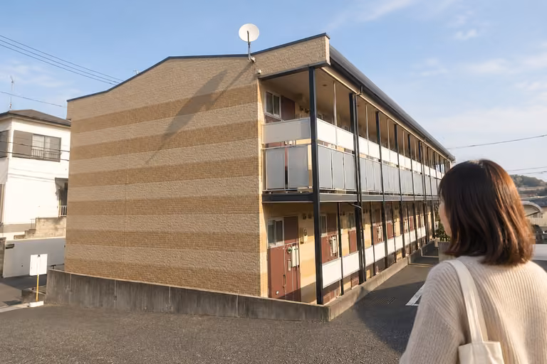 外廊下のある建物を背景に、周囲を確かめる女性の肩越しの様子
