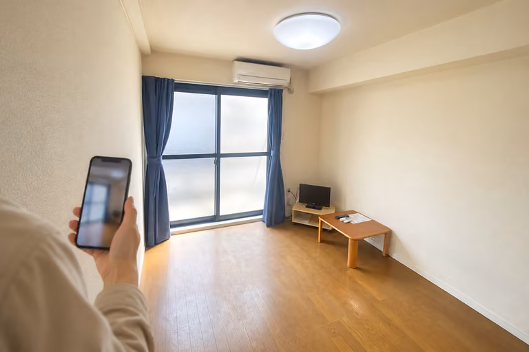 窓とローテーブルのある室内を、スマートフォンで映す肩越しの様子