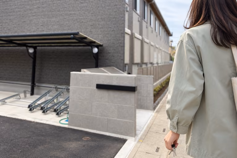 灰色の外壁と駐輪場のある建物へ、鍵を手に帰る女性の後ろ姿